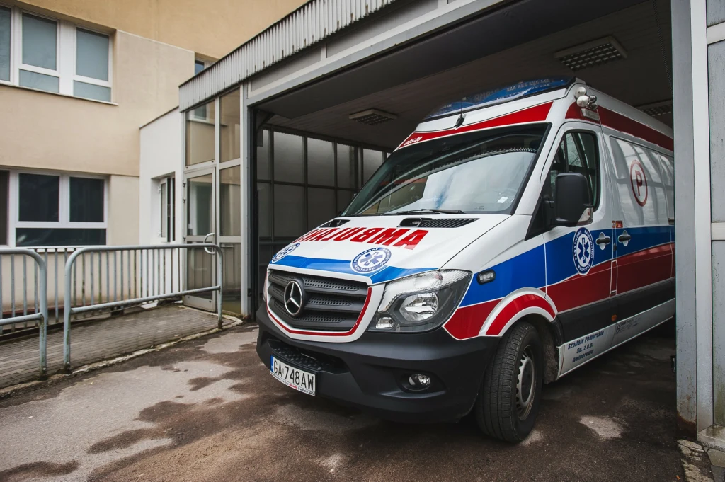 Szef Naczelnej Izby Lekarskiej Łukasz Jankowski: Rozwarstwienie zarobków jest patologiczne Ambulans marki Mercedes zaparkowany przy wejściu do budynku szpitala, częściowo w zadaszonym garażu, z widocznymi oznakowaniami służby ratownictwa medycznego.