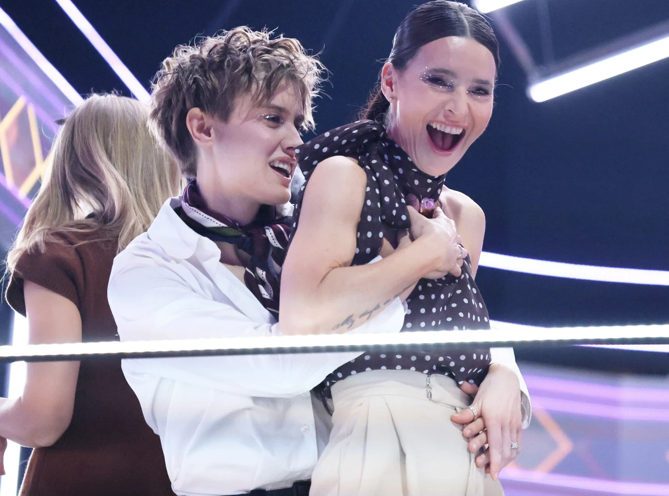 Katarzyna Zillmann i Janja Lesar Dwie uśmiechnięte kobiety w dynamicznej pozie, jedna obejmuje drugą od tyłu, obie wyrażają radość, w tle widoczne abstrakcyjne, jasne światła i geometryczne linie, atmosfera imprezowa lub rozrywkowa.