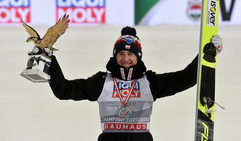 Kamil Stoch ze Złotym Orłem za 69. Turniej Czterech Skoczni Sportowiec w zimowej czapce i medalem na szyi trzyma w jednej ręce dużą statuetkę w kształcie orła, a w drugiej narty. Ubrany w kombinezon z logo sponsorów, na twarzy szeroki uśmiech. W tle rozmazane elementy infrastruktury sportowej.