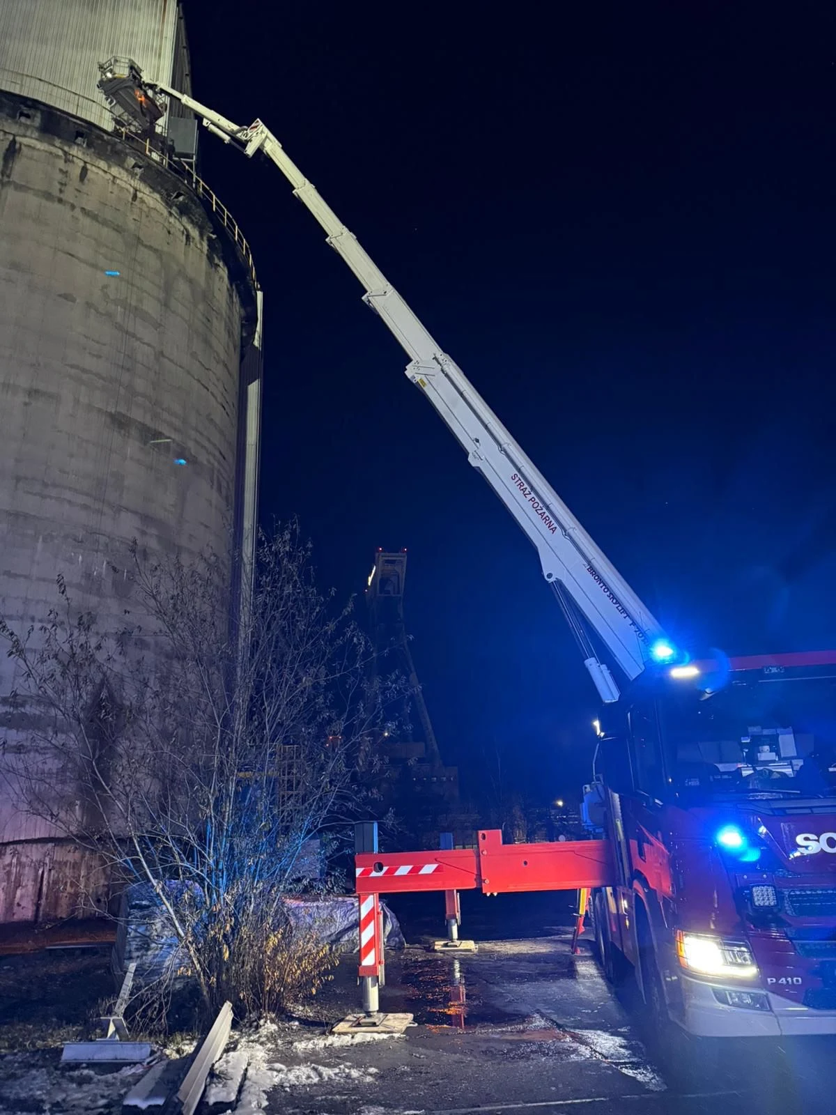 Nocna akcja strażaków przy użyciu dużego podnośnika koszowego skierowanego na szczyt wysokiego, betonowego zbiornika przemysłowego. Pojazd ratowniczy oświetla teren niebieskim światłem, a wokół widać elementy infrastruktury przemysłowej oraz roślinność.