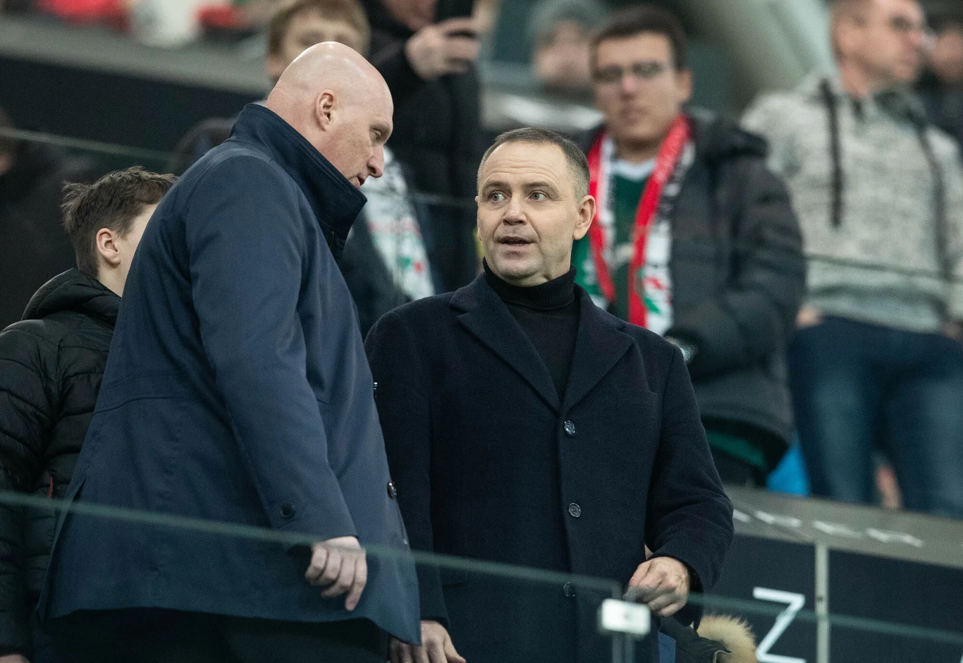 Dwóch mężczyzn w eleganckich płaszczach rozmawia ze sobą na tle trybun stadionu, za nimi widoczni są kibice skupieni na meczu.