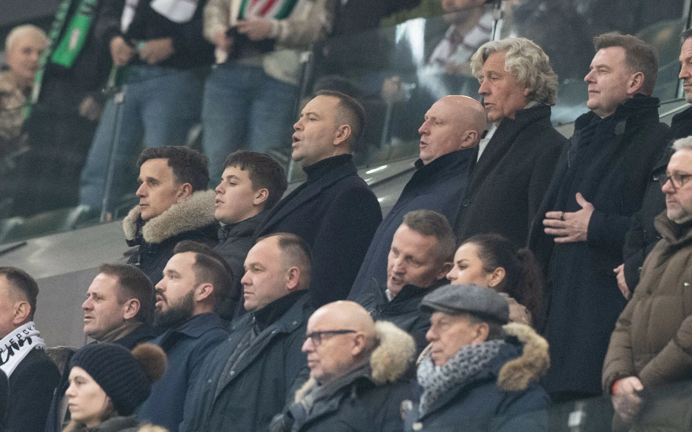 Grupa elegancko ubranych mężczyzn i jedna kobieta stojących na trybunach stadionu, skierowanych w stronę murawy, prawdopodobnie uczestniczących w wydarzeniu sportowym. W tle widać innych kibiców i fragmenty stadionowej infrastruktury.