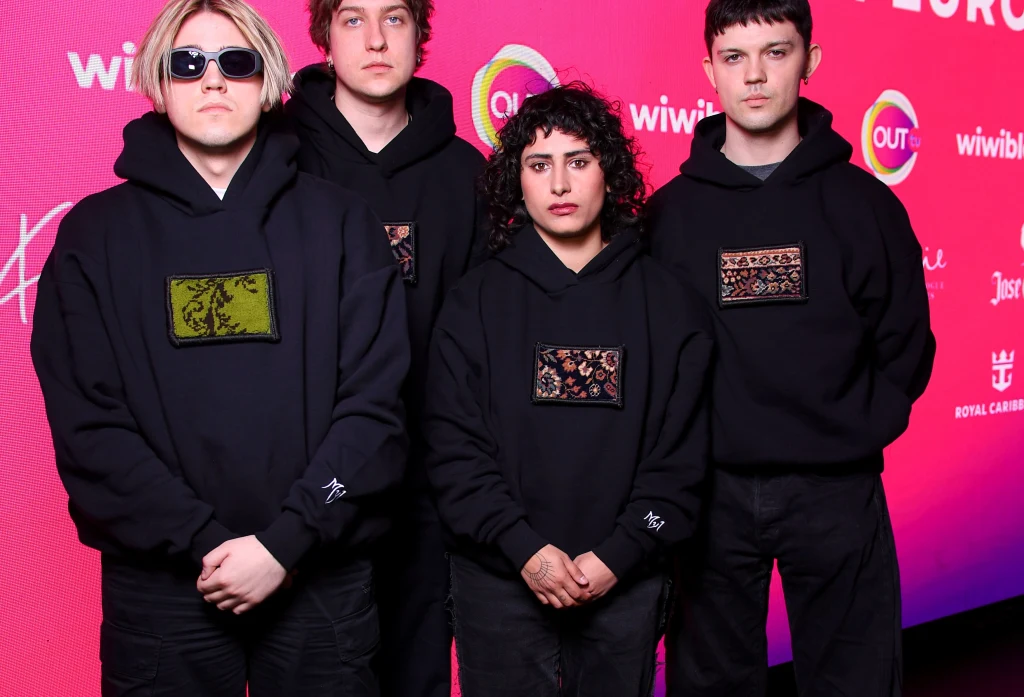 Czworo młodych ludzi ubranych w czarne bluzy z różnymi wzorami na piersi stoi obok siebie na tle różowo-fioletowej ścianki z logo różnych sponsorów i wydarzenia.