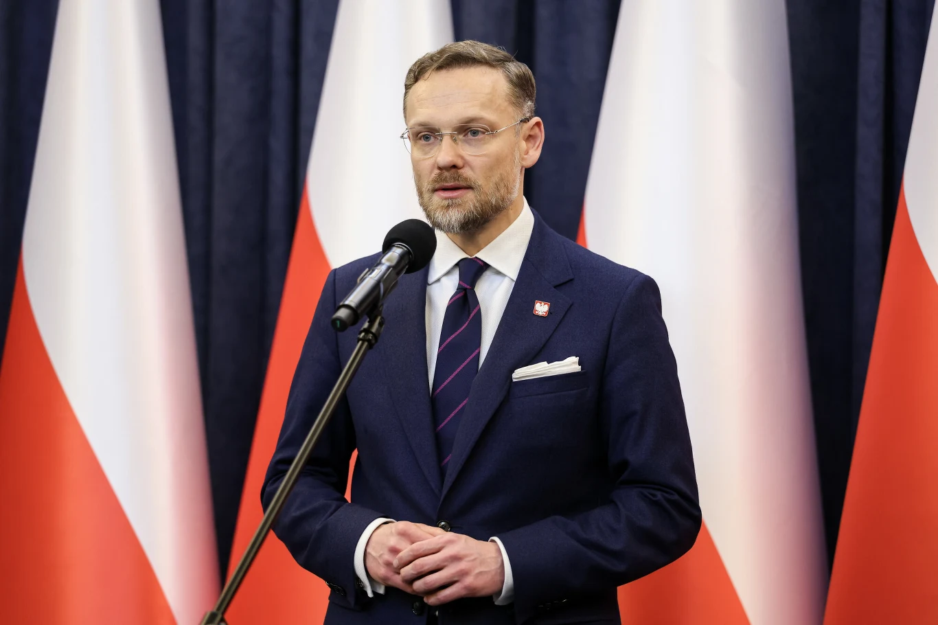 Szef KPRP Zbigniew Bogucki Mężczyzna w garniturze przemawia do mikrofonu, w tle liczne polskie flagi ustawione pionowo.