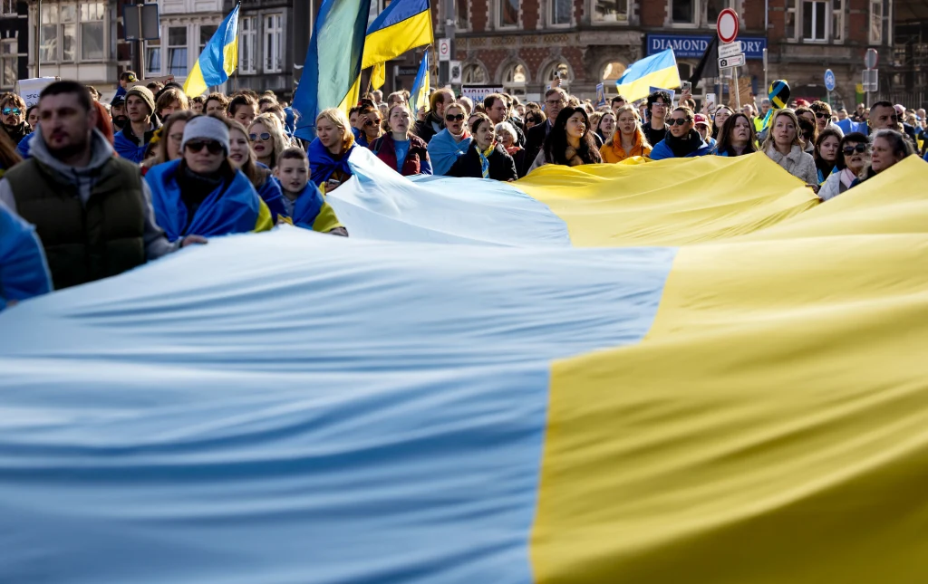 Holandia ma nowe plany wobec Ukraińców, uciekających przed wojną Tłum ludzi maszerujący ulicą trzyma ogromną flagę Ukrainy, większość uczestników ubrana w niebiesko-żółte elementy oraz szaliki, w tle widać miejską zabudowę oraz kolejne flagi ukraińskie.