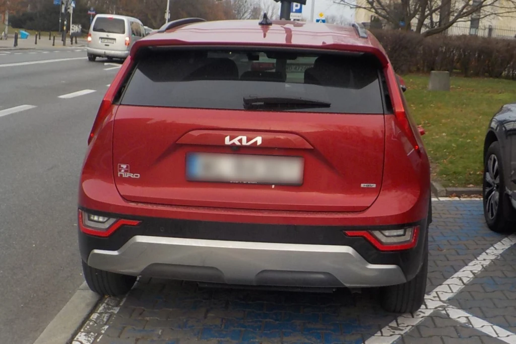 Czerwony samochód marki Kia zaparkowany na miejscu dla osób niepełnosprawnych, fragment innego pojazdu widoczny po prawej stronie, ulica oraz oznaczenia parkingowe w tle.