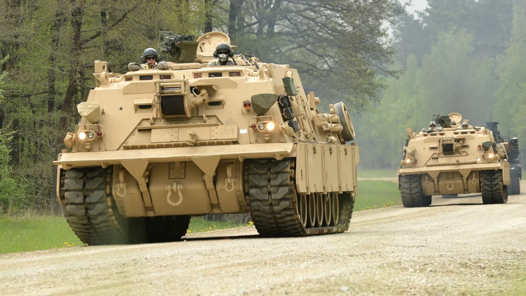 Kolumna wojskowych pojazdów opancerzonych poruszających się po leśnej drodze, w tle widać gęsty las i dwóch żołnierzy znajdujących się na wieżyczkach pojazdów.