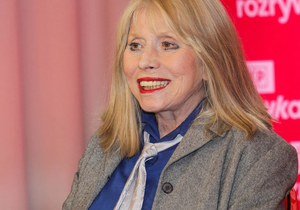 Uśmiechnięta kobieta o blond włosach w szarej marynarce i niebieskiej koszuli z apaszką na tle czerwonego bannera.