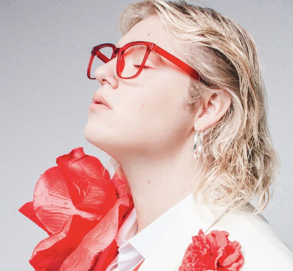 Norbert Wronka prezentuje piosenkę "Boję się" Młoda osoba z blond włosami i dużymi czerwonymi okularami, ubrana w jasny garnitur ozdobiony dużymi czerwonymi kwiatami, oczy zamknięte, twarz skierowana lekko do góry.