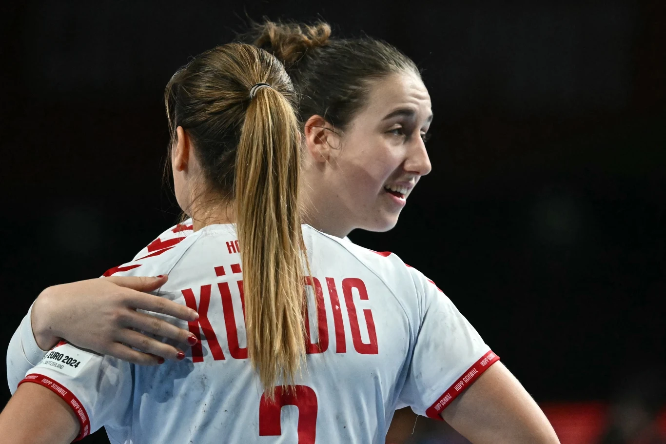 Dwie zawodniczki drużyny piłki ręcznej w białych koszulkach z czerwonymi napisami, jedna obejmuje drugą, panuje sportowa atmosfera wsparcia i koleżeństwa.