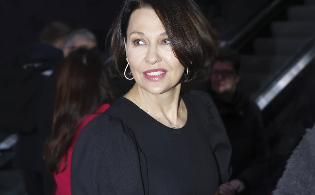 Kobieta o krótkich, ciemnych włosach w eleganckiej, czarnej sukience z okrągłym dekoltem. Zwrócona lekko bokiem, z delikatnym makijażem i kolczykiem w uchu. Tło rozmyte, niewyraźne postacie w tle.