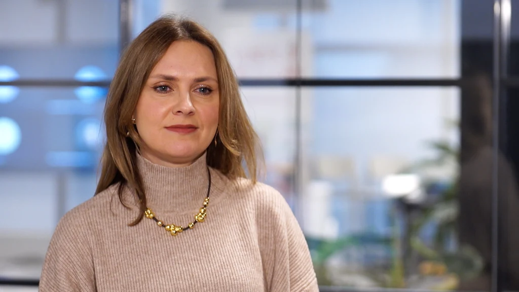 Kobieta o jasnych włosach ubrana w beżowy golf i złoty naszyjnik stoi w nowoczesnym, jasnym pomieszczeniu biurowym z przeszklonymi ścianami w tle.