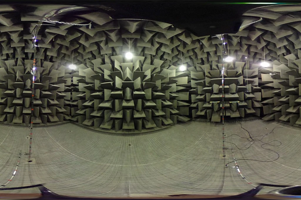 Ściany i sufit pokryte geometrycznymi, stożkowatymi panelami dźwiękochłonnymi, podłoga z siatki, kilka mikrofonów na stojakach oraz przewody, jasne oświetlenie nad konstrukcją.