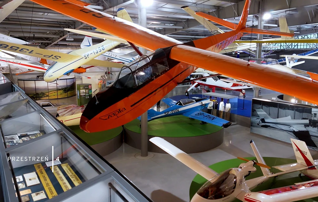 Wnętrze muzeum lotnictwa z licznymi szybowcami i samolotami zawieszonymi pod sufitem, widoczna przeszklona ekspozycja oraz plansze informacyjne, dominuje duży pomarańczowy szybowiec na pierwszym planie.