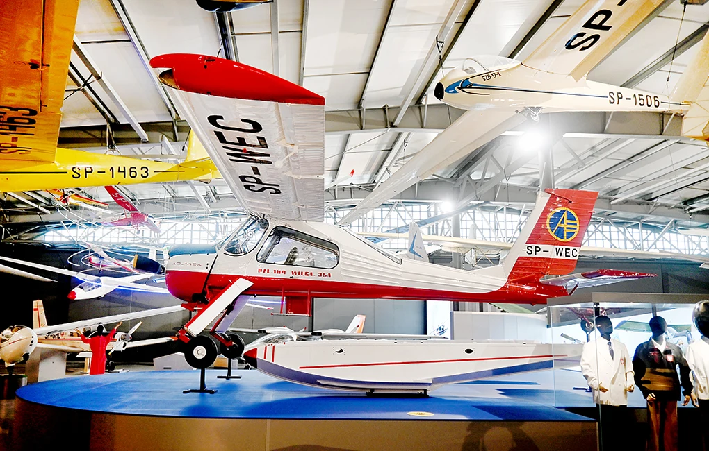 Samolot sportowy z biało-czerwonym kadłubem oraz oznaczeniami SP-WEC ustawiony na ekspozycji w muzeum lotnictwa. W tle widoczne inne samoloty oraz elementy hali wystawowej.