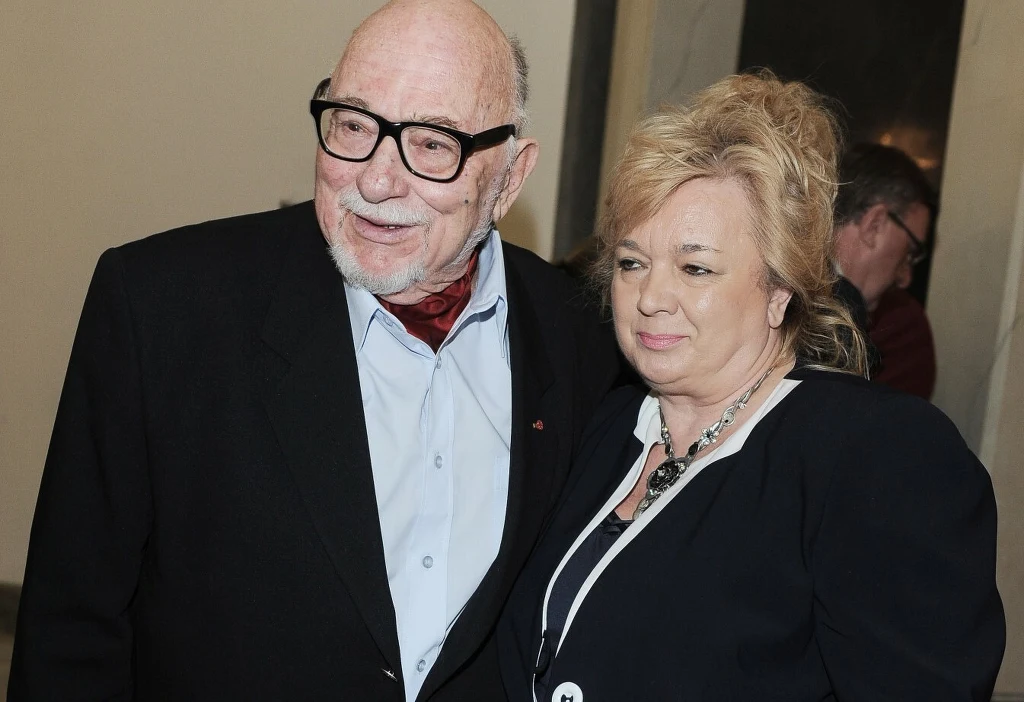 Jerzy Hoffman z żoną Jagodą Prądzyńską Starszy mężczyzna w ciemnym garniturze, białej koszuli i czerwonej apaszce oraz kobieta w ciemnej marynarce z jasnymi włosami stoi obok siebie, oboje uśmiechnięci, tworząc wrażenie bliskości i ciepłej relacji.