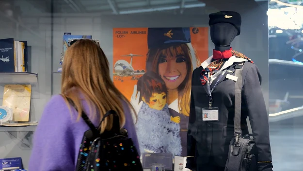 Młoda kobieta z plecakiem ogląda wystawę z manekinem ubranym w mundur stewardesy, obok widoczny plakat Polskich Linii Lotniczych LOT z uśmiechniętą stewardesą trzymającą dziecko, w tle różne eksponaty związane z lotnictwem.