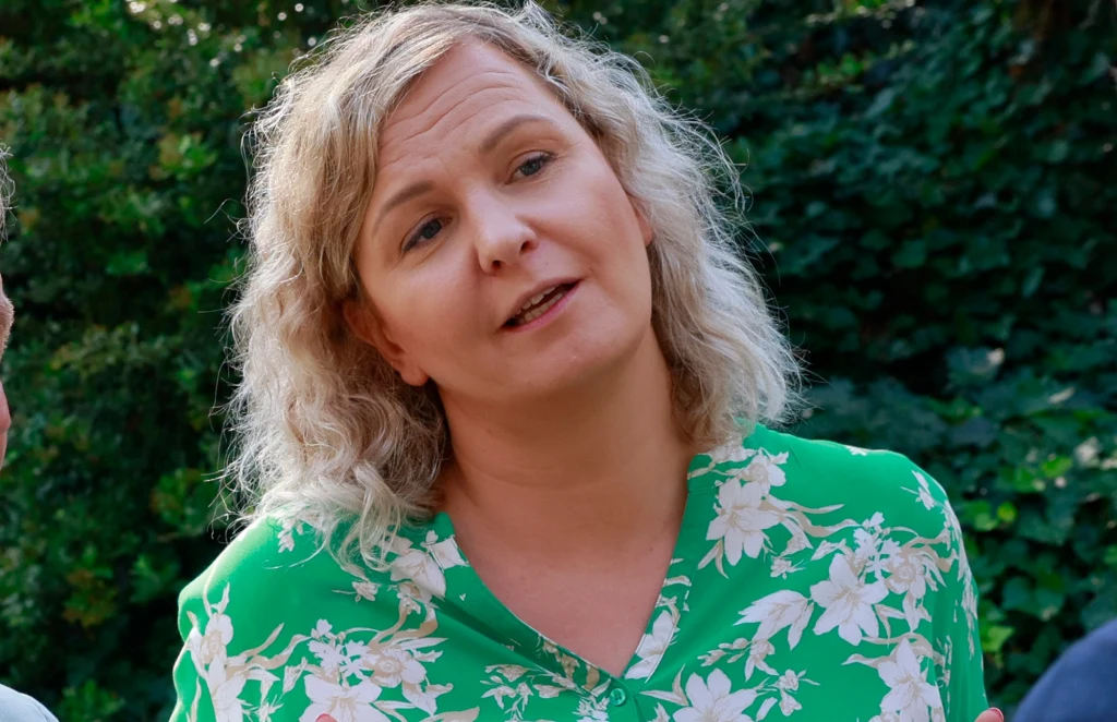 Otylia Jędrzejczak Kobieta o blond kręconych włosach w zielonej bluzce w kwiatowy wzór stoi na tle zieleni, jej wyraz twarzy jest spokojny i zamyślony.