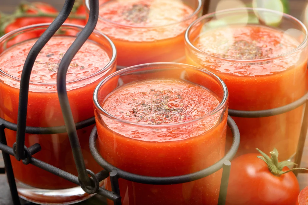 Na co jest dobry sok pomidorowy? Lista właściwości jest długa Szklanki wypełnione świeżo przygotowanym sokiem pomidorowym, posypane czarnym pieprzem na wierzchu, ustawione w metalowym koszyku. W tle widoczne całe pomidory.