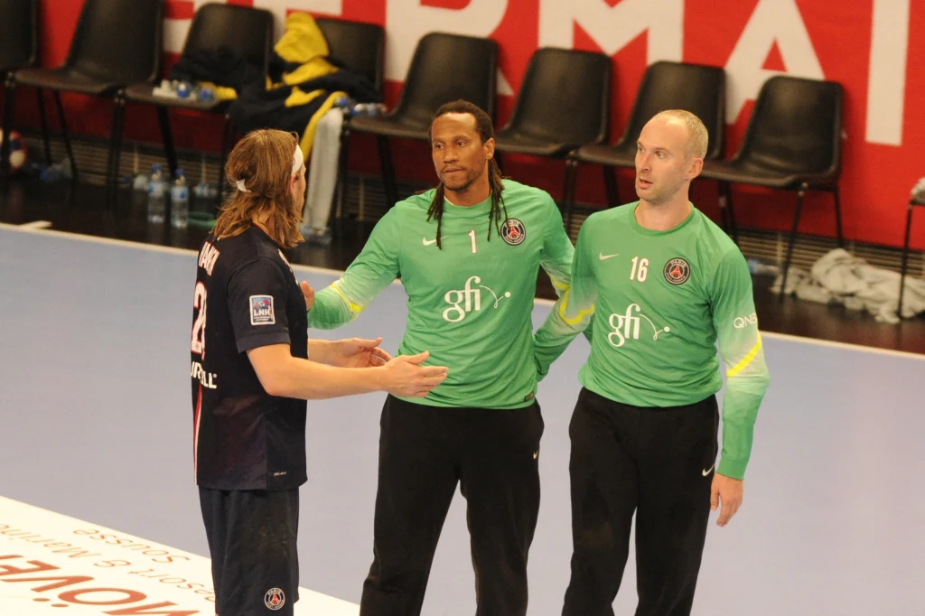 Od lewej: Mikkel Hansen, Patrice Annonay i Thierry Omeyer Dwóch zawodników w zielonych koszulkach rozmawia z innym sportowcem w ciemnym stroju na hali sportowej, w tle rząd pustych krzeseł i fragment dużego czerwonego napisu na ścianie.