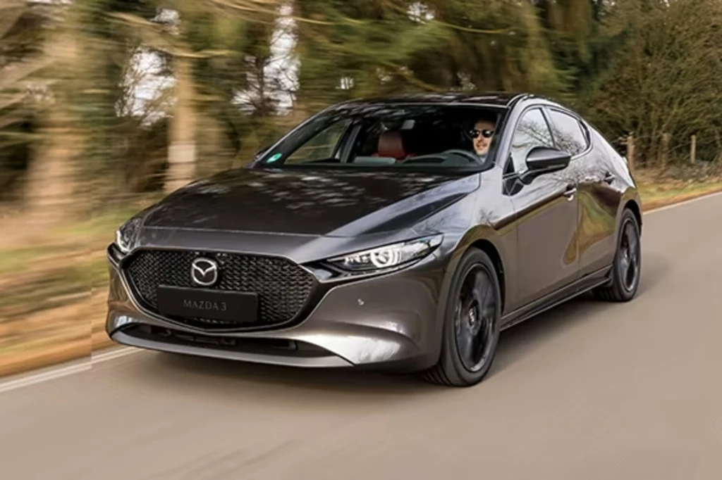 Silnik SKYACTIV-X znajdziemy m.in. pod maską Mazdy 3 Czarny samochód osobowy marki Mazda 3 jedzie asfaltową drogą przez zalesiony teren, za kierownicą siedzi kierowca w okularach przeciwsłonecznych.
