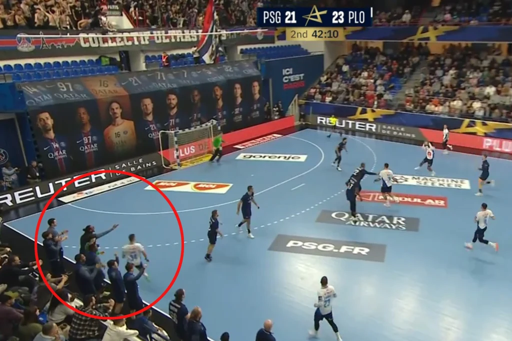 To tu Lovro Mihić "nadział się" na rękę Patrice'a Annonaya Mecz piłki ręcznej rozgrywany na profesjonalnej hali sportowej, widoczne drużyny PSG oraz PLO. Tablica wyników pokazuje wynik 21:23 oraz czas gry 42:10. Po lewej stronie w pobliżu ławki rezerwowych zawodnicy oraz członkowie sztabu technicznego podejmuj...