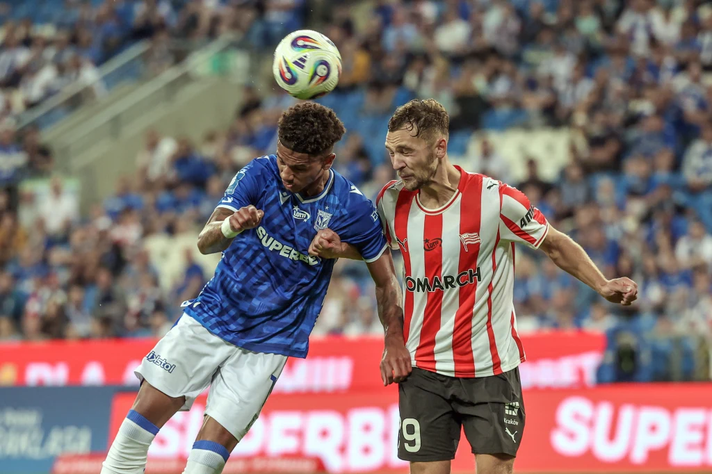 Dwóch piłkarzy walczy o piłkę w powietrzu podczas meczu piłki nożnej na stadionie pełnym kibiców, jeden z drużyny w niebieskich barwach, drugi w biało-czerwonych paskach.
