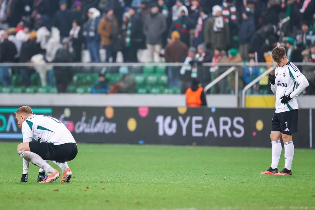 Niepocieszeni piłkarze Legii Dwóch piłkarzy w białych strojach z opuszczonymi głowami na murawie stadionu, wokół trybuny pełne kibiców. Sportowa atmosfera i emocje po zakończonym spotkaniu.