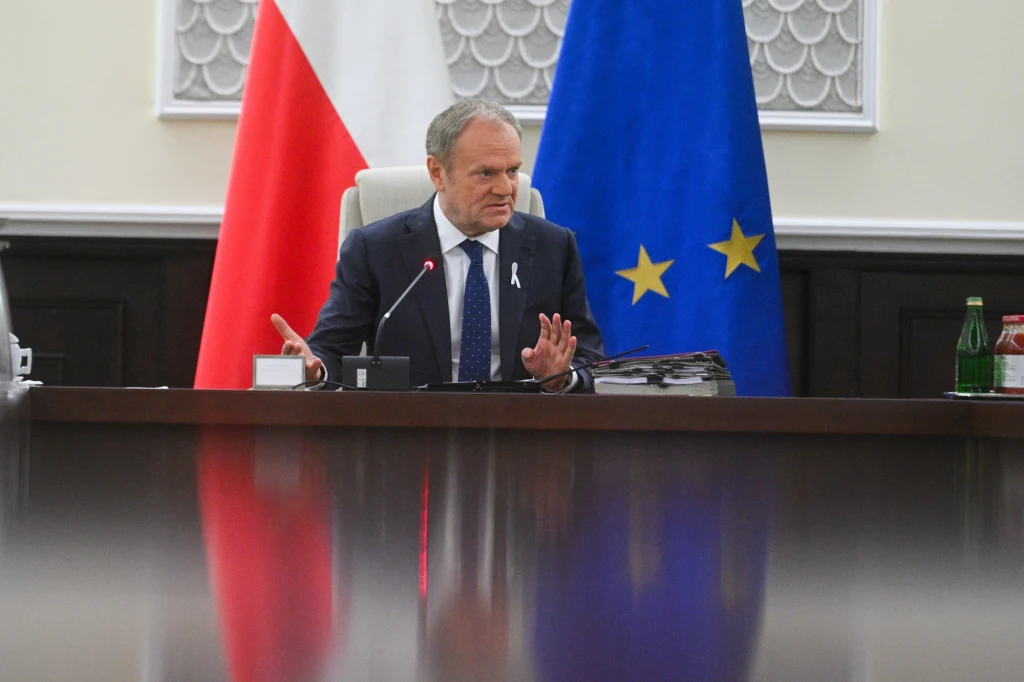 Premier Donald Tusk odniósł się do sprawy orzeczenia TSUE podczas ostatniego posiedzenia rządu Mężczyzna w garniturze siedzi przy dużym, drewnianym stole konferencyjnym, w tle widoczne są flagi Polski oraz Unii Europejskiej. Na stole znajdują się dokumenty, mikrofon oraz butelka wody.
