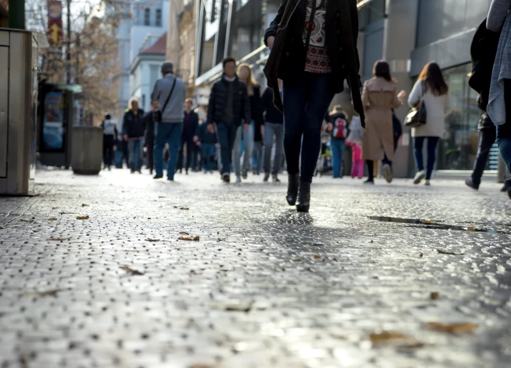 Ulica miasta pokryta kamiennym brukiem z grupą przechodniów, z których większość idzie w kierunku aparatu, a w tle widoczne są nowoczesne budynki i drzewa ozdobione jesiennymi liśćmi.