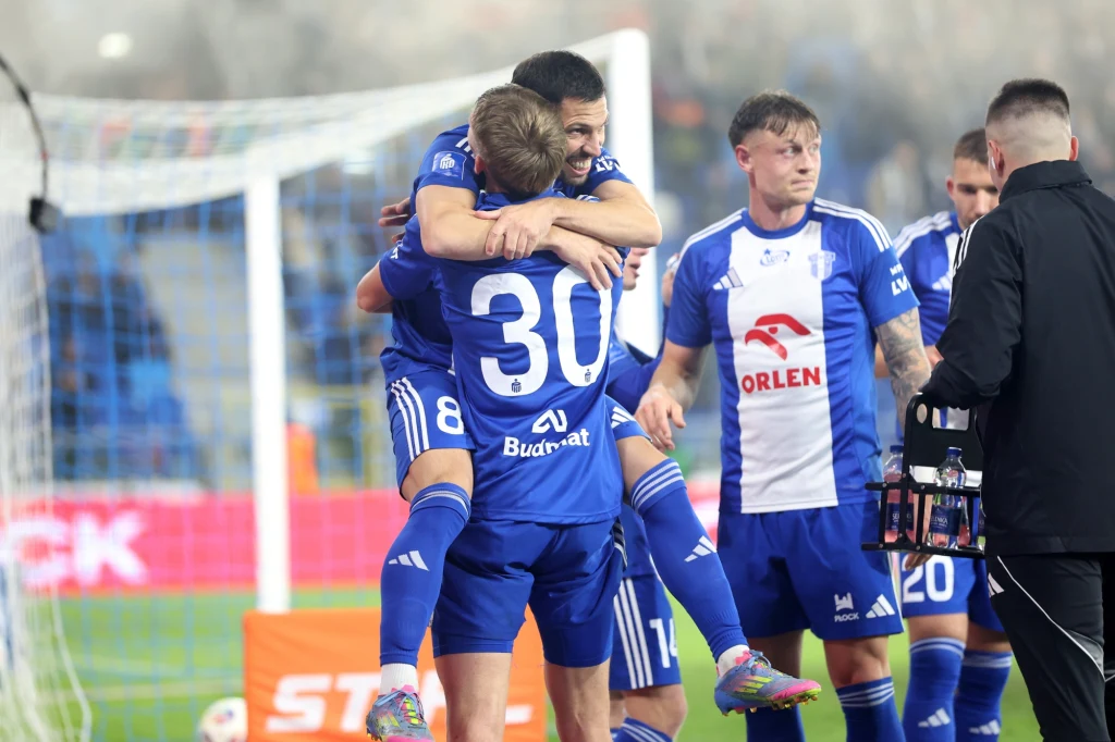 Piłkarze w niebieskich strojach świętują zdobycie bramki, jeden z zawodników podnosi drugiego, pozostali gracze obserwują celebrację na tle trybun i innych członków drużyny.