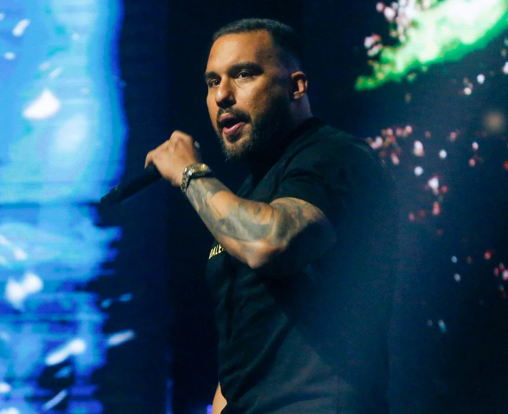 Jongmen jest dobrze znany na scenie hiphopowej Mężczyzna z tatuażem na przedramieniu trzymający mikrofon podczas występu na scenie, dynamiczne oświetlenie w niebiesko-zielonych barwach w tle.