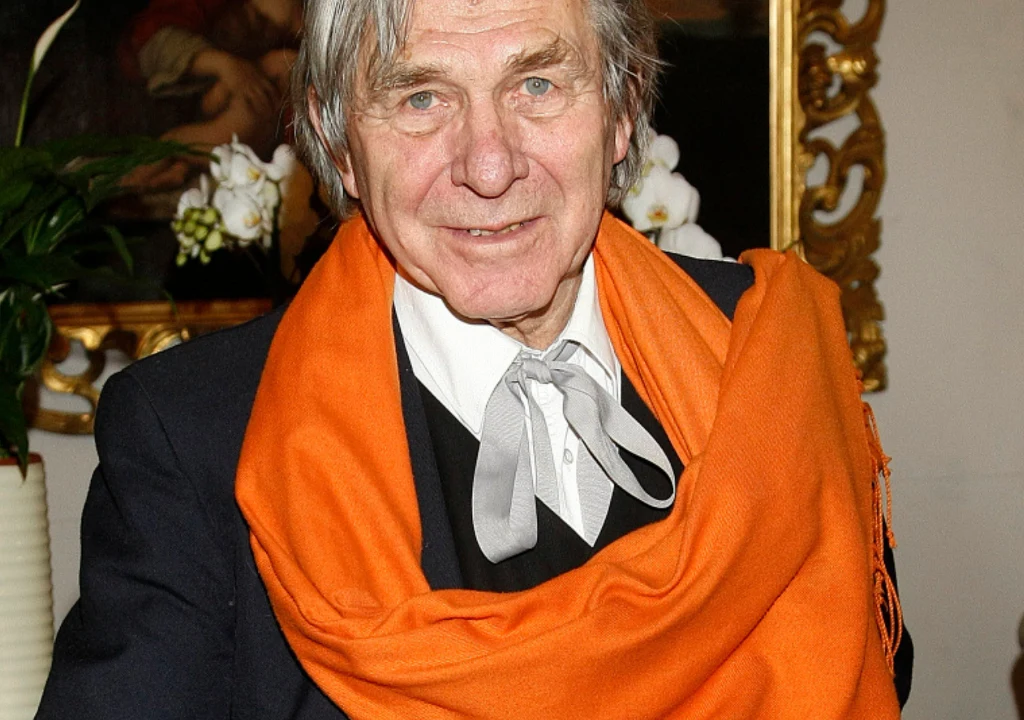 Starszy mężczyzna z siwymi włosami ubrany w ciemny garnitur z białą koszulą, szarym krawatem i pomarańczowym szalem, w tle ozdobne ramy oraz kwiaty.