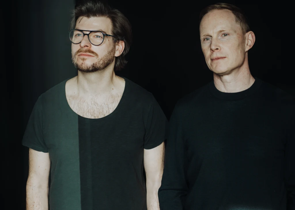 Salamon & Fitzsimons Dwóch mężczyzn stoi obok siebie na ciemnym tle, jeden z nich ma okulary i zarost, a obaj noszą czarne koszulki.