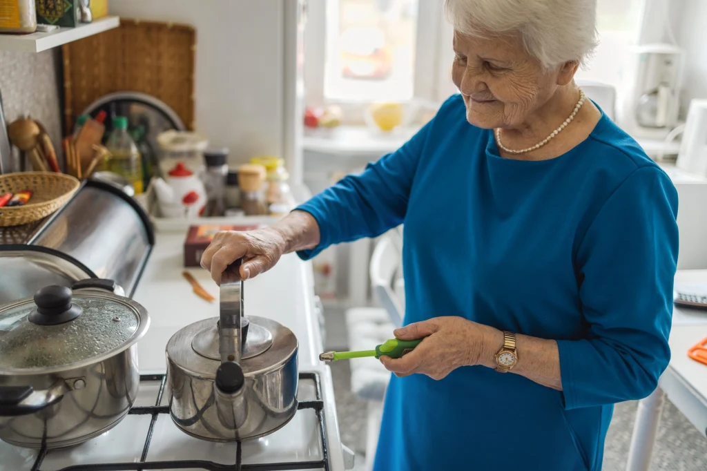 Starsza kobieta w niebieskiej sukience stoi w kuchni przy kuchence gazowej, trzyma garnek i zapalniczkę w rękach. W tle widoczne są kuchenne przybory i przyprawy.