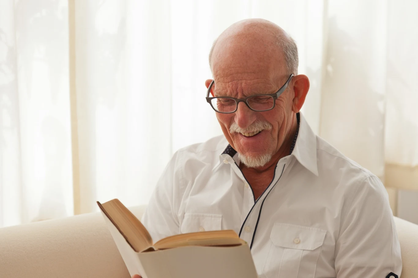 Starszy mężczyzna w okularach siedzi na kanapie i z uśmiechem czyta książkę, ubrany w białą koszulę, w tle jasne zasłony przepuszczające światło dzienne.
