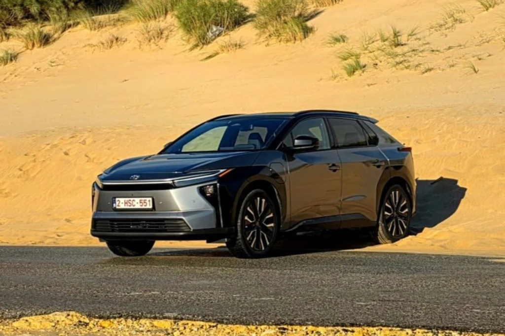 Czarny samochód osobowy marki Toyota stoi zaparkowany na poboczu asfaltowej drogi na tle żółtego piasku i roślinności na wydmach.