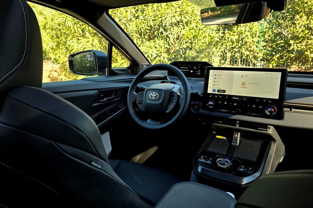 Nowoczesne wnętrze samochodu Toyota z dużym dotykowym ekranem centralnym, wielofunkcyjną kierownicą i czarnymi skórzanymi fotelami. Poza samochodem widoczna zieleń i naturalne światło wpadające przez okna.