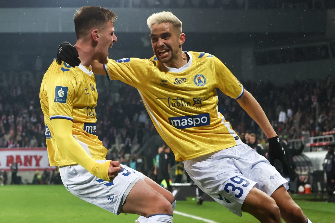 Dwaj piłkarze drużyny w żółtych koszulkach celebrują zdobycie bramki na stadionie pełnym kibiców, wyrażając radość i sportowe emocje.