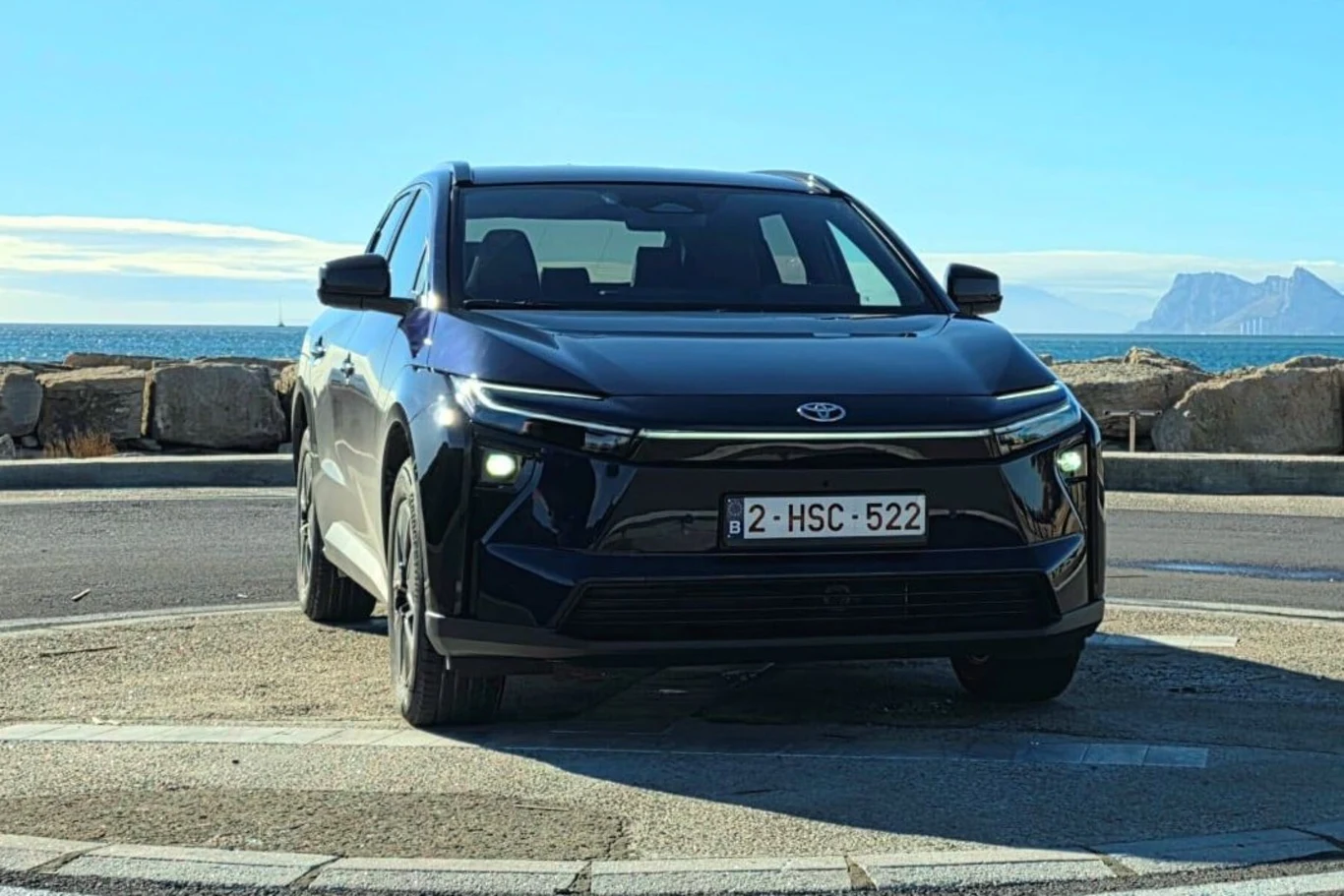 Czarny samochód marki Toyota zaparkowany na tle nadmorskiego pejzażu z widocznymi skałami i górami w oddali, przy słonecznej pogodzie.