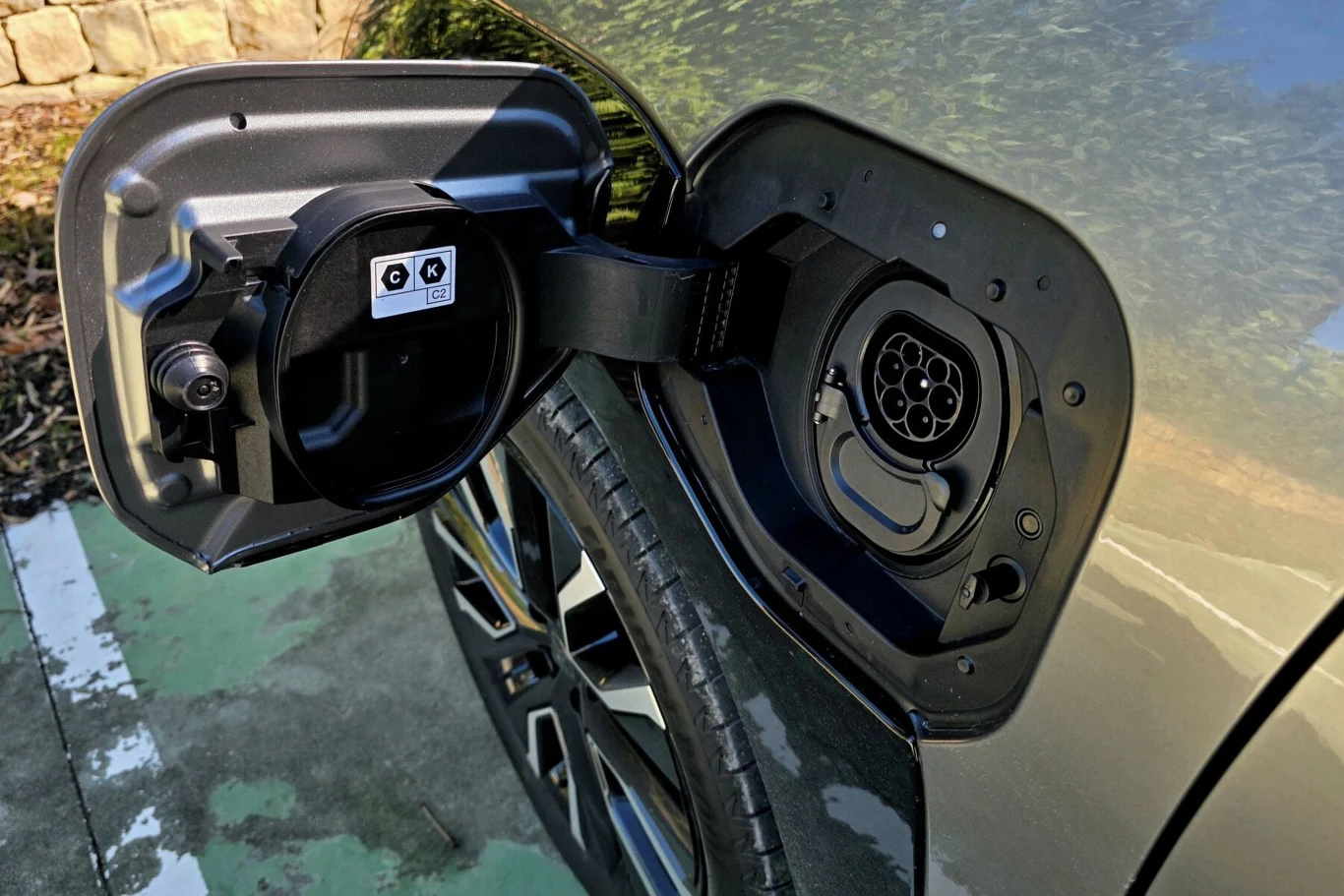 Port ładowania samochodu elektrycznego otwarty na tle fragmentu koła oraz nadwozia pojazdu, widoczne oznaczenia techniczne na pokrywie, auto stoi na parkingu z zielonym oznaczeniem świadczącym o miejscu ładowania.