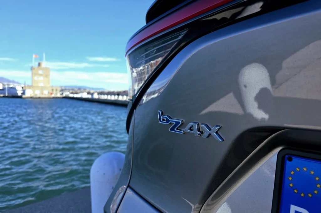 Tylny fragment srebrnego samochodu elektrycznego Toyota bZ4X zaparkowanego przy nabrzeżu, na tle wody i budynku portowego; widoczne oznaczenie modelu oraz fragment europejskiej tablicy rejestracyjnej.