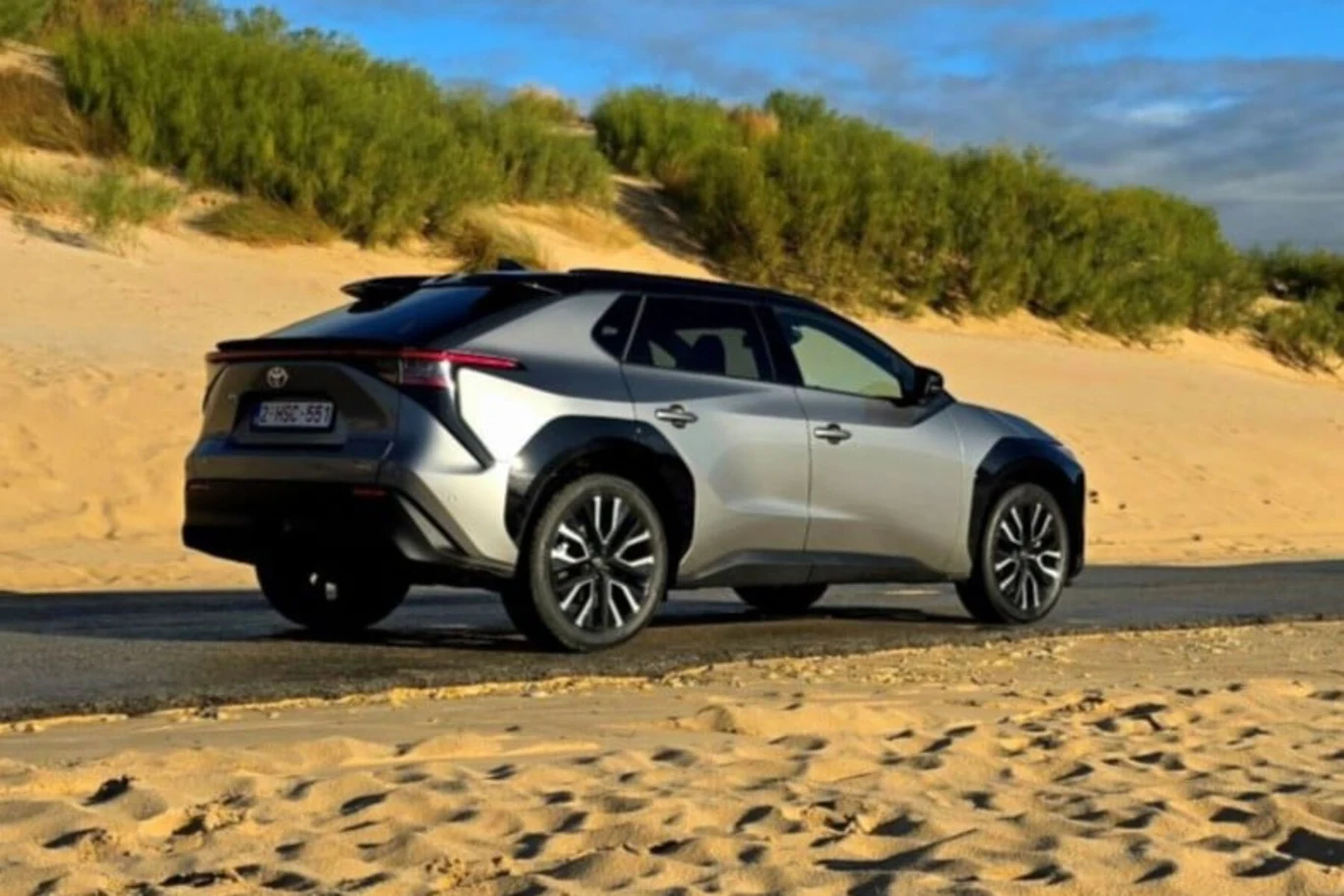 Srebrny samochód terenowy marki Toyota stoi zaparkowany na asfaltowej drodze przy piaszczystych wydmach z zieloną roślinnością w tle, pod niebieskim niebem z rozproszonymi chmurami.