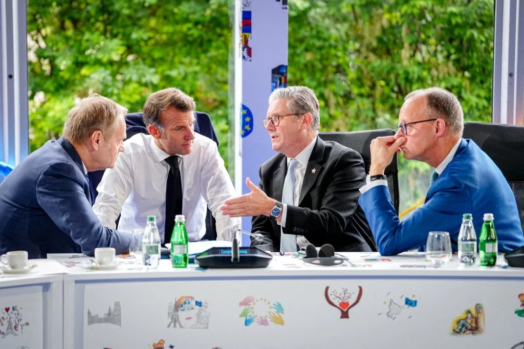 Czterech mężczyzn w garniturach siedzi przy stole, prowadząc poważną rozmowę. Na stole znajdują się butelki z wodą, filiżanki i dokumenty, a w tle widoczna jest zieleń zza okna.