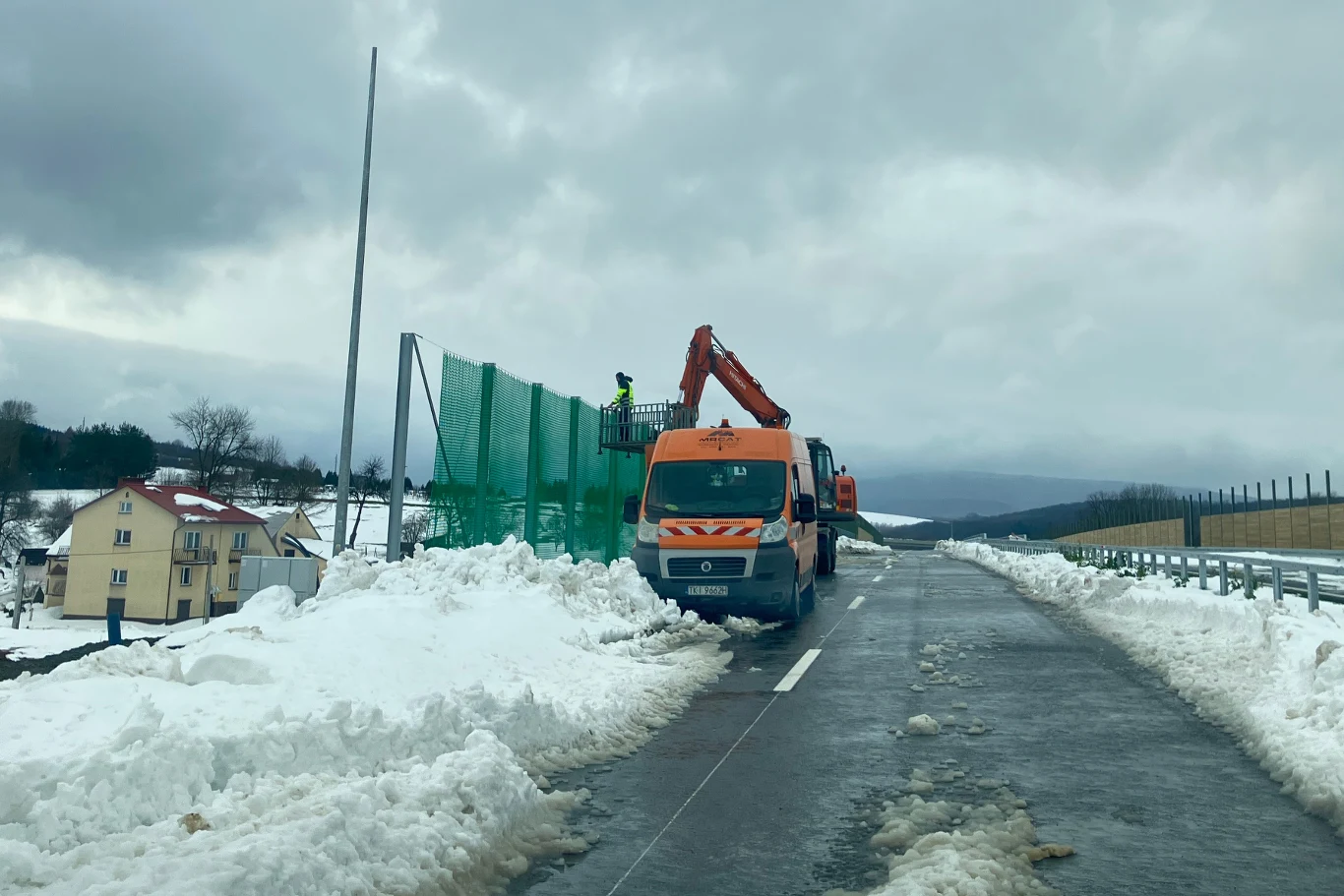 GDDKiA montuje 4-metrowe siatki przy drodze S19. Zaśnieżona droga szybkiego ruchu, po której porusza się pomarańczowy samochód serwisowy. Obok pojazdu znajduje się dźwig z osłoną akustyczną oraz pracownicy w kamizelkach odblaskowych. Na poboczu zalega śnieg, w tle domy i wzgórza.