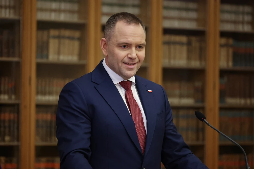 Mężczyzna w granatowym garniturze i czerwonym krawacie stoi przy mównicy, za nim znajdują się regały z książkami, co nadaje scenie poważny i oficjalny charakter.