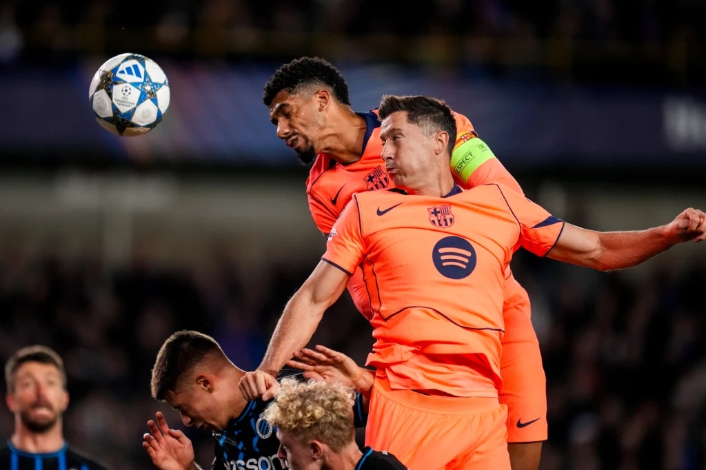 Ronald Araujo i Robert Lewandowski Dwóch piłkarzy w pomarańczowych strojach FC Barcelony wyskakuje do główki podczas meczu piłkarskiego, walcząc o piłkę z przeciwnikami. Jeden z zawodników trzyma opaskę kapitańską, w tle widoczni inni gracze i rozmyta publiczność stadionowa.