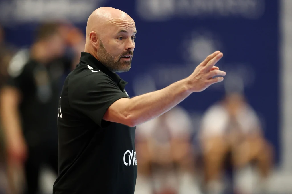 Łysy mężczyzna w czarnej koszulce gestykuluje ręką, prawdopodobnie podczas prowadzenia drużyny sportowej, w tle rozmyci zawodnicy i niebieskie tło z niewyraźnym napisem.