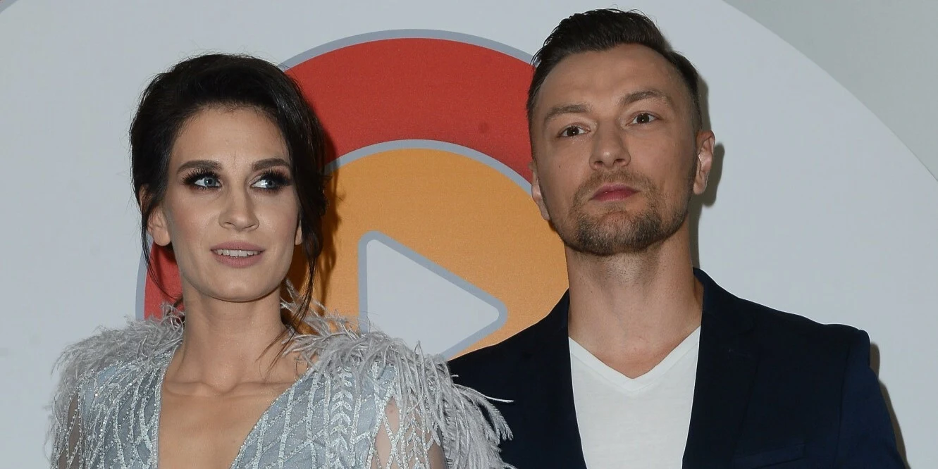 Sylwia Grzeszczak i Liber do 2023 r. byli parą Kobieta w jasnej, wieczorowej sukience z ozdobnymi piórami stoi obok mężczyzny w ciemnej marynarce i białym t-shircie na tle logo z dużym przyciskiem play.