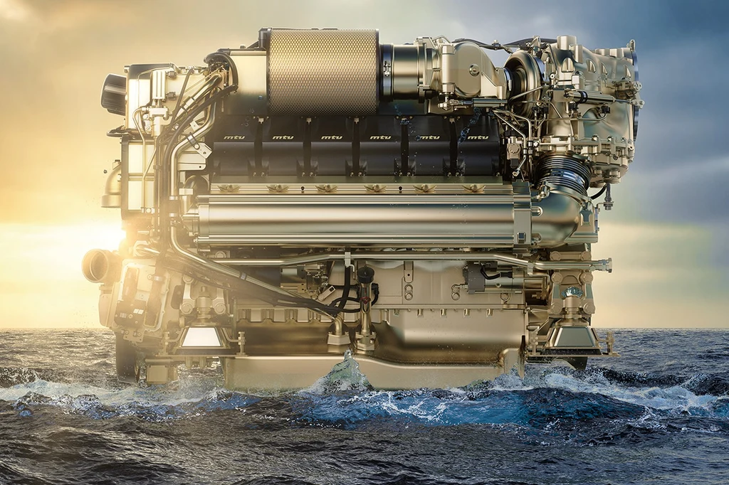 Duży silnik wysokoprężny unoszący się nad powierzchnią morza, oświetlony światłem zachodzącego słońca. Woda rozpryskuje się wokół silnika, tło stanowi jasne, lekko zachmurzone niebo.